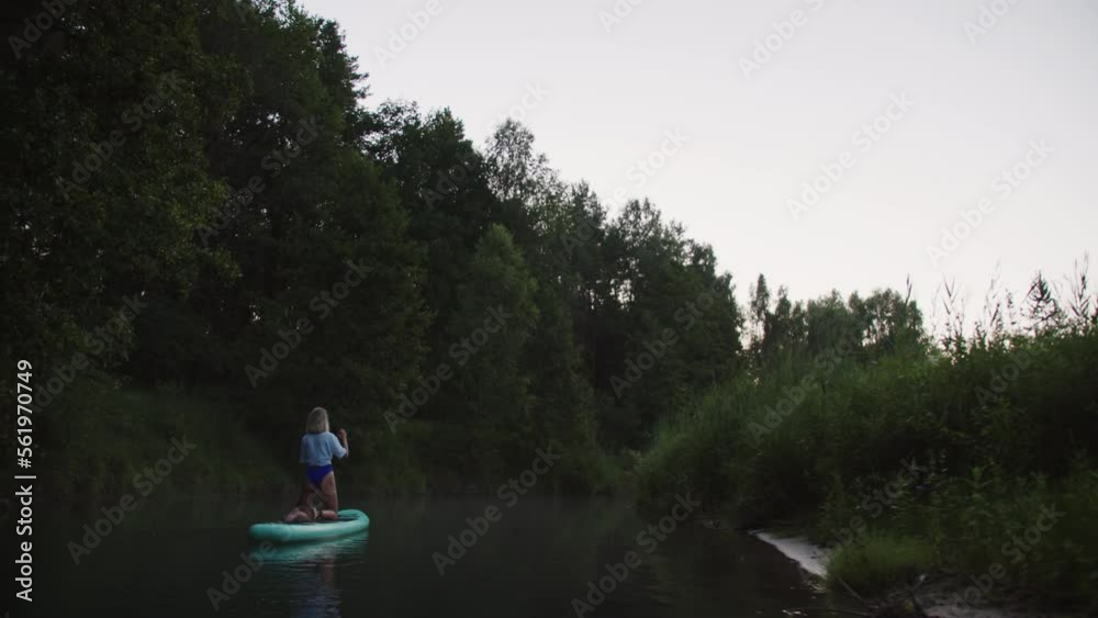 Young woman paddling on surfing board, standing on knees, man swimming after sportswoman rowing on paddleboard, front view. Female coach rows on surfboard. Sports lesson for beginners on water. Young woman paddling on surfing board, standing on knees, man swimming after sportswoman rowing on paddleboard, front view. Female coach rows on surfboard. Sports lesson for beginners on water.