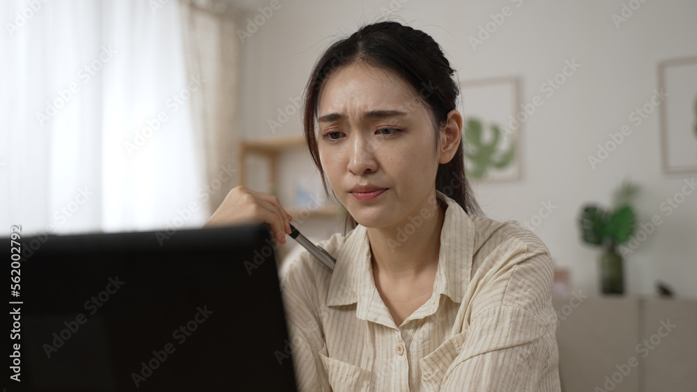 frustrated asian businesswoman troubled by a problem while working from home is propping head and staring at computer screen thinking hard about solutions. frustrated asian businesswoman troubled by a problem while working from home is propping head and staring at computer screen thinking hard about solutions.