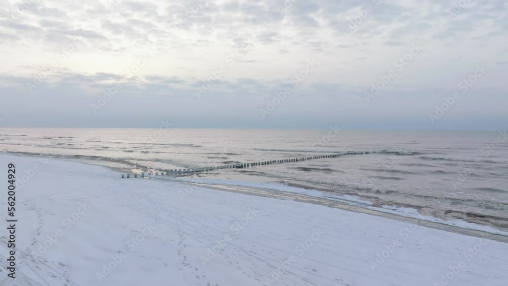 Aerial establishing view of an old wooden pier at the Baltic sea coastline, overcast winter day, white sand beach covered in snow, ice on wood poles, calm seashore, wide drone shot moving forward Aerial establishing view of an old wooden pier at the Baltic sea coastline, overcast winter day, white sand beach covered in snow, ice on wood poles, calm seashore, wide drone shot moving forward