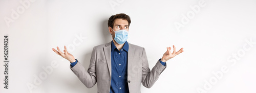 Confused businessman in medical mask shrugging and looking aside, know nothing, cant understand something, standing on white background photo