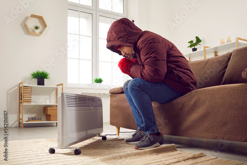 Warmly dressed man warming hands at electric heater feeling cold at home. Man sitting on sofa wearing warm jacket and gloves and warming himself at cold winter season © Studio Romantic
