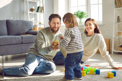 Cute little girl playing game with her parents at home. Smiling dad giving golden Christmas ball to his toddler daughter, mom looking at her with tenderness. Happy family playing toys together