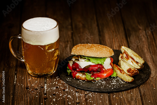 Homemade beef burger with mug of cold beer, baked potato, tomatoes, cheese, ketchup on wood table 