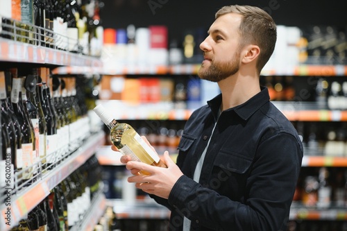Fotografie, Obraz Portrait of young glad positive male customer selecting wine in supermarket