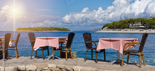 Take a seat, set table of a restaurant on the Croatian Adriatic, on a stone wall, directly above the blue, crystal clear sea. In the background green overgrown islands and cloudy sky.