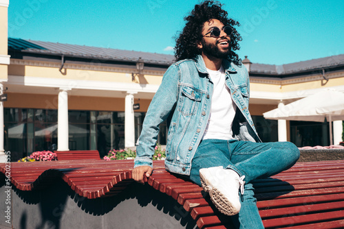 Handsome smiling hipster model. Sexy unshaven Arabian man dressed in summer jeans jacket clothes. Fashion male with long curly hairstyle posing in the street at sunset. In sunglasses