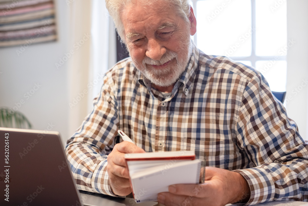 Smiling mature bearded man sitting at table with laptop and books following online course. Senior man in checkered shirt enjoying teaching activity and learning using new technology Smiling mature bearded man sitting at table with laptop and books following online course. Senior man in checkered shirt enjoying teaching activity and learning using new technology