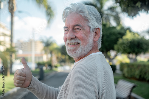 Portrait of cheerful optimist senior man looking at camera with thums up. Elderly bearded caucasian male in outdoor in urban park enjoying retirement photo