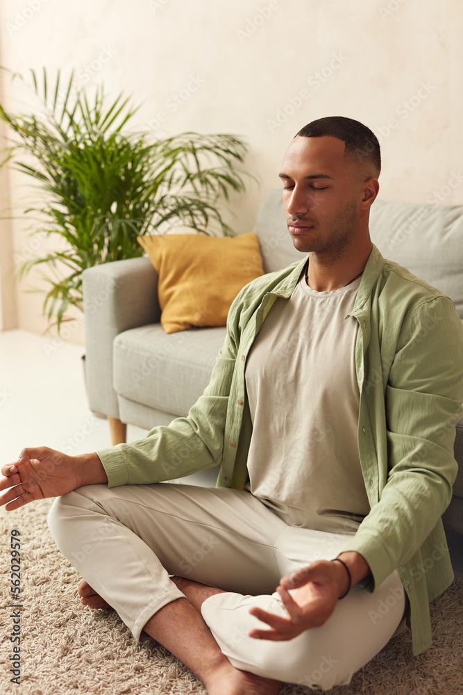Fototapeta premium Calm Man Meditating Home. Multiracial Guy Sitting In Lotus Pose Practicing Yoga At Home. Sport, Fitness And Healthy Lifestyle Concept 