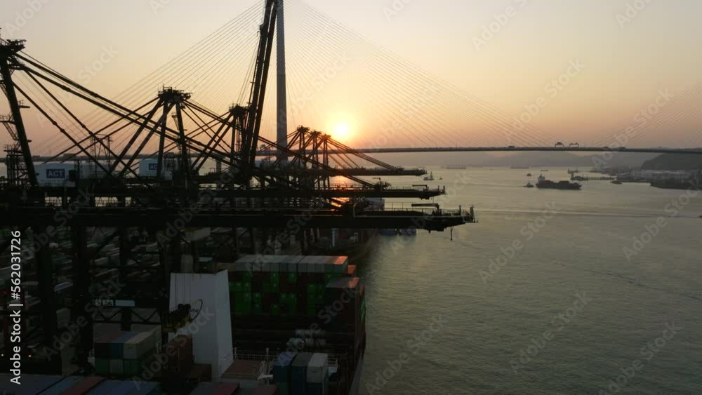 Gantry cranes loading and discharging containers on moored ships with a large suspension bridge in background during sunset. Drone lifting shot Gantry cranes loading and discharging containers on moored ships with a large suspension bridge in background during sunset. Drone lifting shot