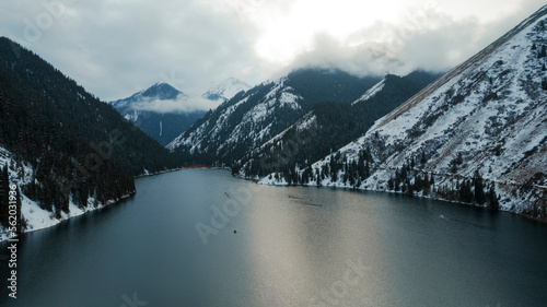 Kolsai mountain lake in the winter forest. Drone view of clouds, coniferous trees, mirrored smooth water, hills and mountains in the snow. Yellow sunset. Boats float in places. Kazakhstan, Almaty