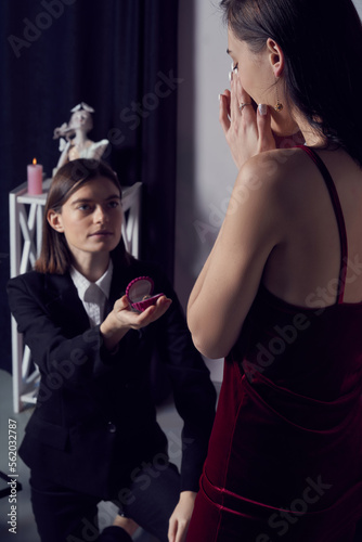 Cropped close-up shot of a brunette girl in black suit giving a ring in a box to her girlfriend. Lady in a red dress excited to receive the meaningful gift from her sweetheart. LGBT concept.
