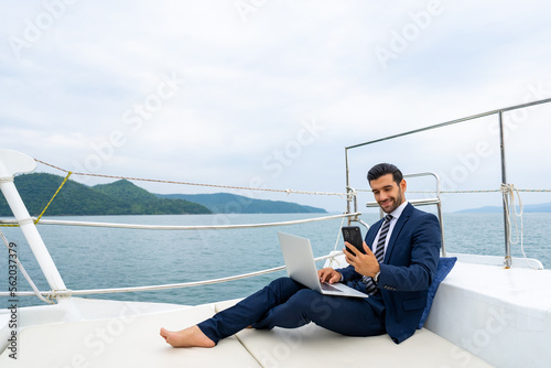 Caucasian businessman working on laptop computer and talking on mobile phone for global corporate business while travel on luxury private catamaran boat yacht sailing in the ocean on summer vacation. © CandyRetriever 