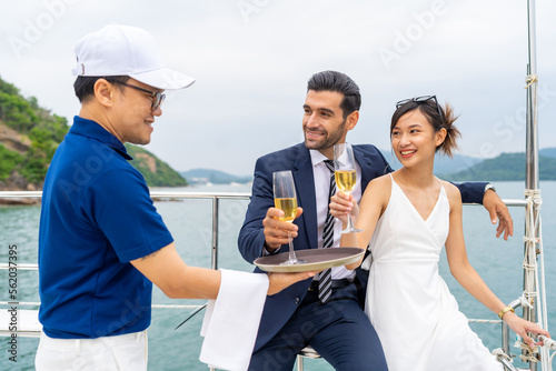 Asian man waiter serving champagne to passenger tourist while private catamaran boat yacht sailing in the ocean. Happy couple enjoy luxury outdoor lifestyle travel on summer beach holiday vacation.