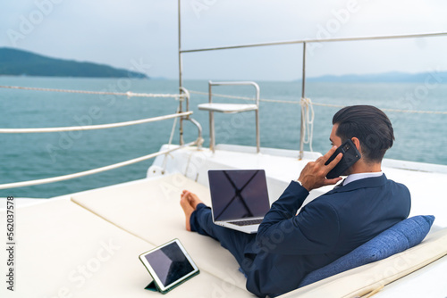 Foto Caucasian businessman working on laptop computer and talking on mobile phone for global corporate business while travel on luxury private catamaran boat yacht sailing in the ocean on summer vacation