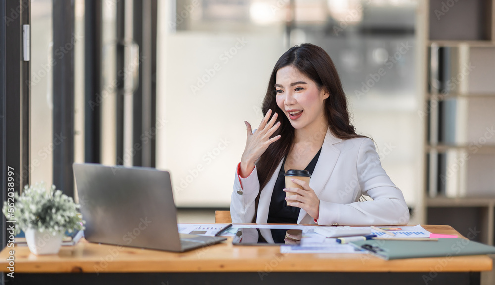 Asian Businesswoman having a tea, sitting on desk in workplace, writing down notes, opened laptop in document the financial report, business plan investment, finance analysis concept.