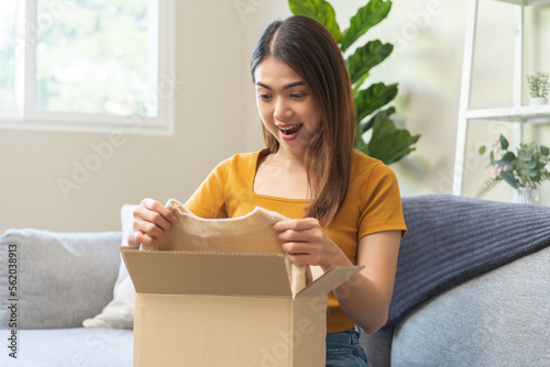 Happy excited, asian young woman, girl customer sitting on sofa at home, open and unpack cardboard box carton parcel after buy fashion clothes, ordering present and shopping online, delivery service.