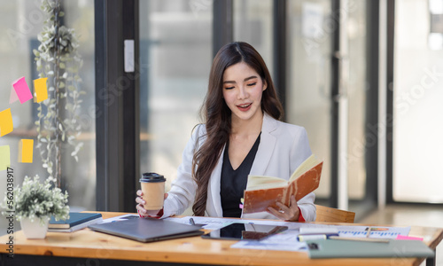 Asian Businesswoman having a tea, sitting on desk in workplace, writing down notes, opened laptop in document the financial report, business plan investment, finance analysis concept. © David