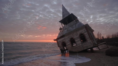 Mysterious vintage flooded leaning wooden house or chapel washed by sea waves, at coastline on pink sunset. Enigmatic rickety building sinking in water on sandy beach near breakwater on sunrise. photo