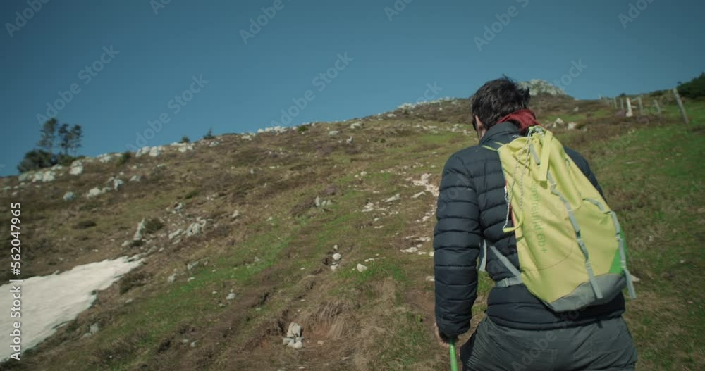 Camera tracking a male hiker with a light green backpack and green hiking poles on a uphill path on the mountain Ratitovec. Clear sky. Camera tracking a male hiker with a light green backpack and green hiking poles on a uphill path on the mountain Ratitovec. Clear sky.