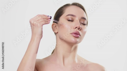 Big close-up shot of attractive European young woman applies face care serum on her cheek from dropper on white background. Face care serum advertising