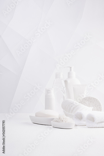 Modern minimalist soft light white bathroom with cosmetic product and toiletry - towels  bath salt  sponge on white table for body and skin care  in contemporary geometric style  copy space  vertical.