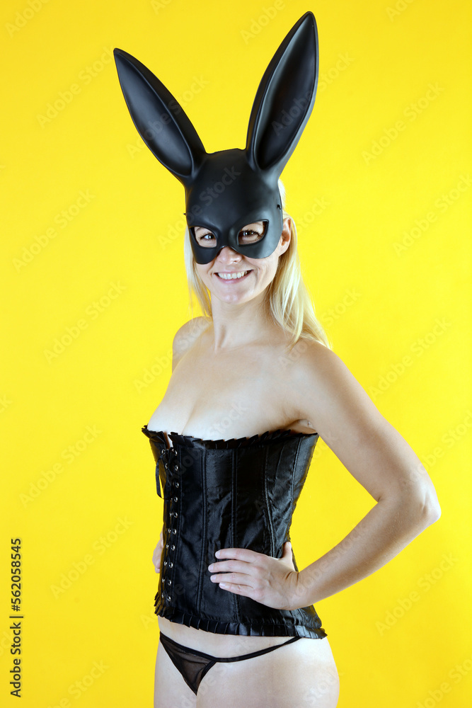 Beautiful blonde woman wears mask with bunny ears for eastern, posing in black corsage in front of yellow background in studio Beautiful blonde woman wears mask with bunny ears for eastern, posing in black corsage in front of yellow background in studio