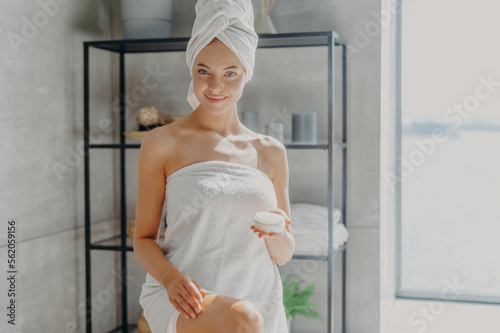 Horizontal shot of pleased smiling woman wrapped in bath towel, touches legs, has silky skin, uses cosmetic product after taking shower, has epilation procedure. Female applies moisturising cream