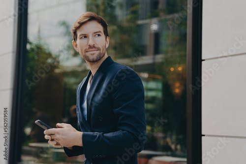 Successful handsome businessman messaging to client on smartphone while standing next to building