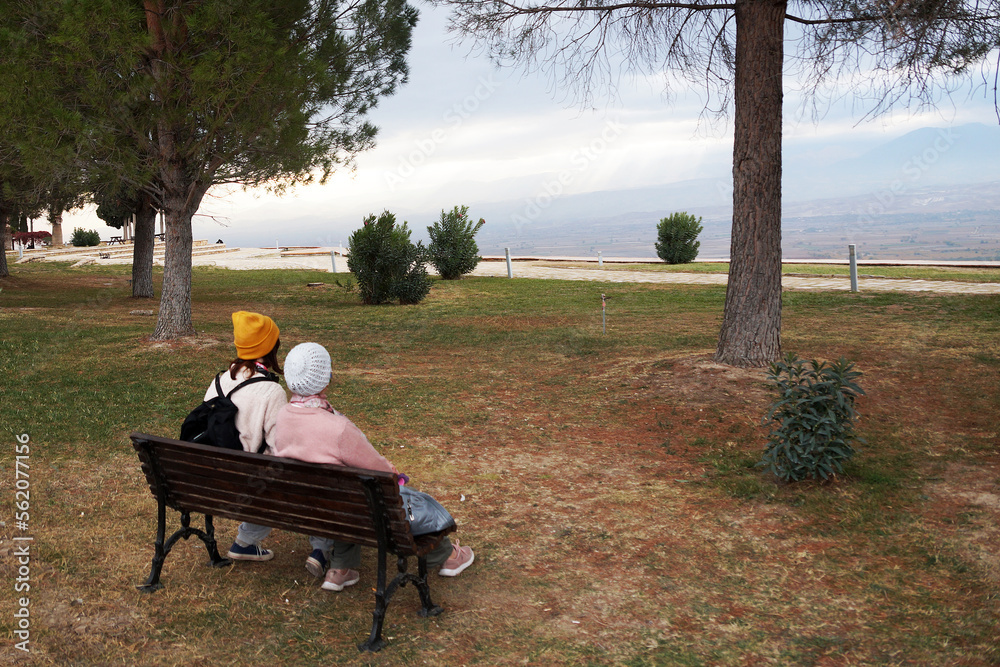 granddaughter and grandmother sit on a bench in the park and look at the mountain panorama granddaughter and grandmother sit on a bench in the park and look at the mountain panorama