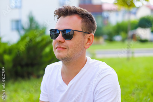 Young male man portrait. European caucasian white young student in sunglasses on green grass lawn on town street, park. Lifestyle Headshot portrait, tourist, businessman