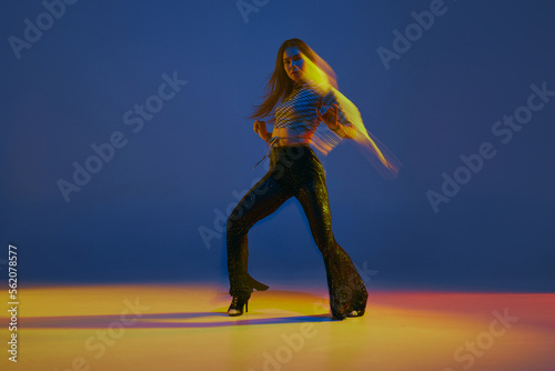 Moxed lights. Portrait of young girl dancing heels dance in stylish clothes over blue background in neon light. Concept of modern dance style
