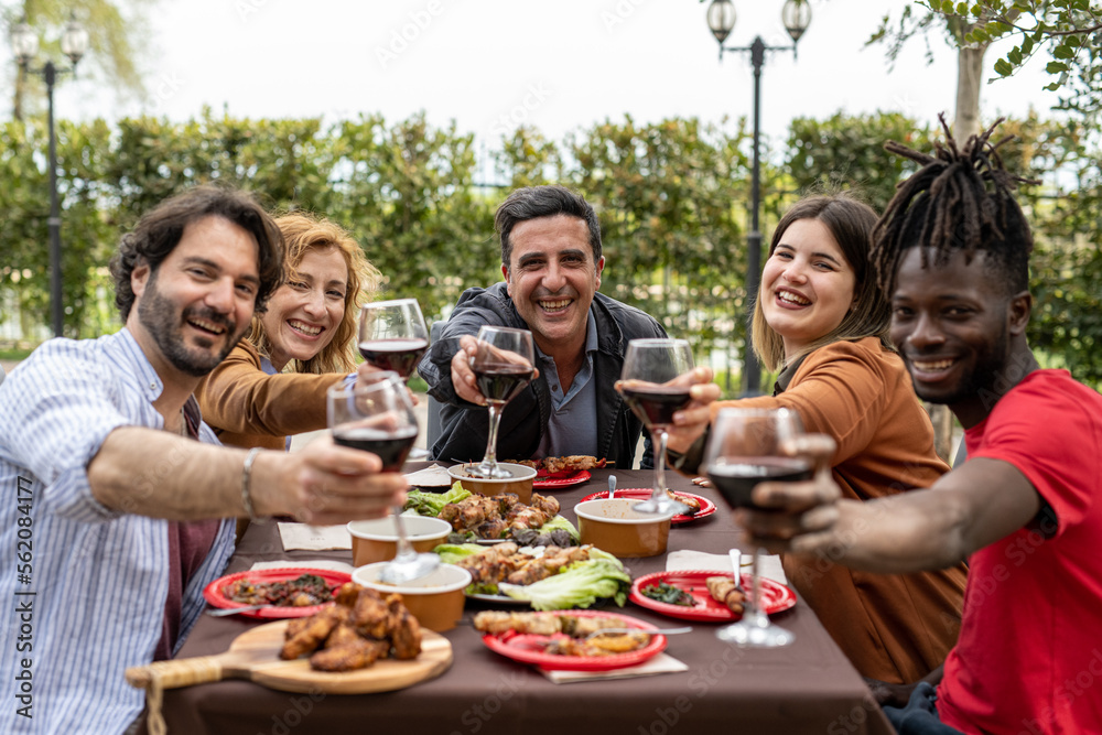 Obraz premium Happy trendy family cheering with red wine at barbecue lunch outdoor, different age of people having fun at sunday meal, food, taste and unity concept, focus on mature man sitting at the head