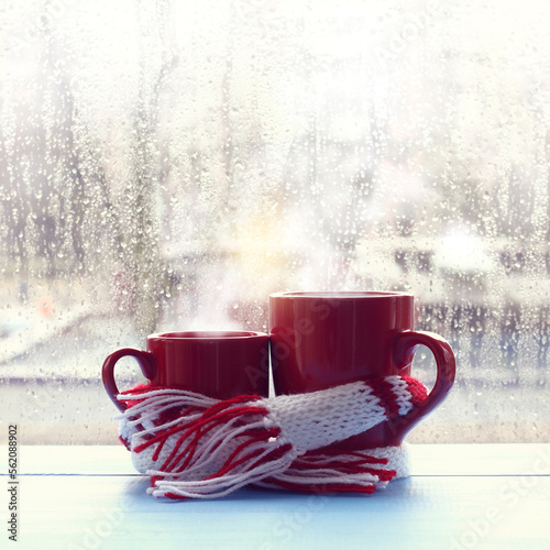 small and large red mugs are wrapped in a scarf against the background of a window with raindrops. together in the warm morning atmosphere