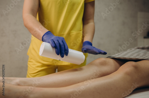 Cosmetologist is sprinkling and distributing talcum powder on a young girl's leg before the epilation procedure. The girl is lying on a couch in a beauty salon, she does the procedure shugaring