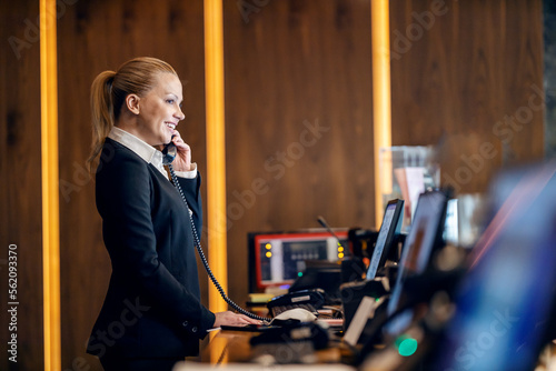 Fotografia A happy receptionist is talking on the phone with hotel guest while standing at reception