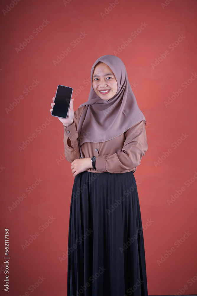 Beautiful Asian woman in brown shirt and hijab smiling cheerful has smartphone with black screen in hand, pointing with index finger at product on brown background Beautiful Asian woman in brown shirt and hijab smiling cheerful has smartphone with black screen in hand, pointing with index finger at product on brown background