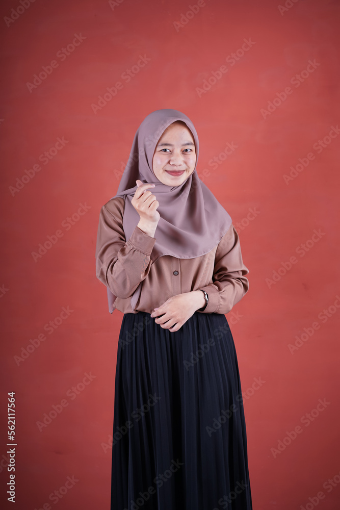  Beautiful Asian woman in brown shirt and hijab smiling cheerfully showing Korean heart with two fingers crossed, express joy and positivity on brown background