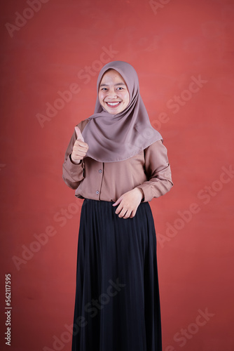 Beautiful Asian woman in brown shirt and hijab smiling cheerfully shows upset expression with thumbs up on brown background
