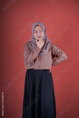  Beautiful Asian woman in brown shirt and hijab showing thinking expression with hand on chin on brown background