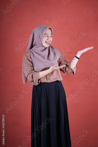 Beautiful Asian woman in brown shirt and hijab smiling cheerful presenting and pointing with palm of hand on brown background 