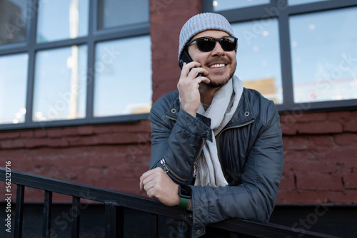 a man in sunglasses and a hat with a scarf speaks with a smile on the phone outside