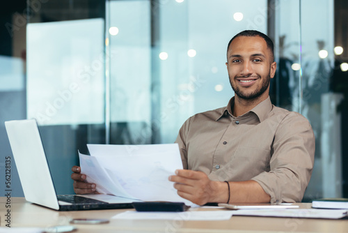 Photographie Portrait of young successful hispanic businessman inside office, man smiling and looking at camera, paper worker happy with achievement results sitting at workplace with laptop