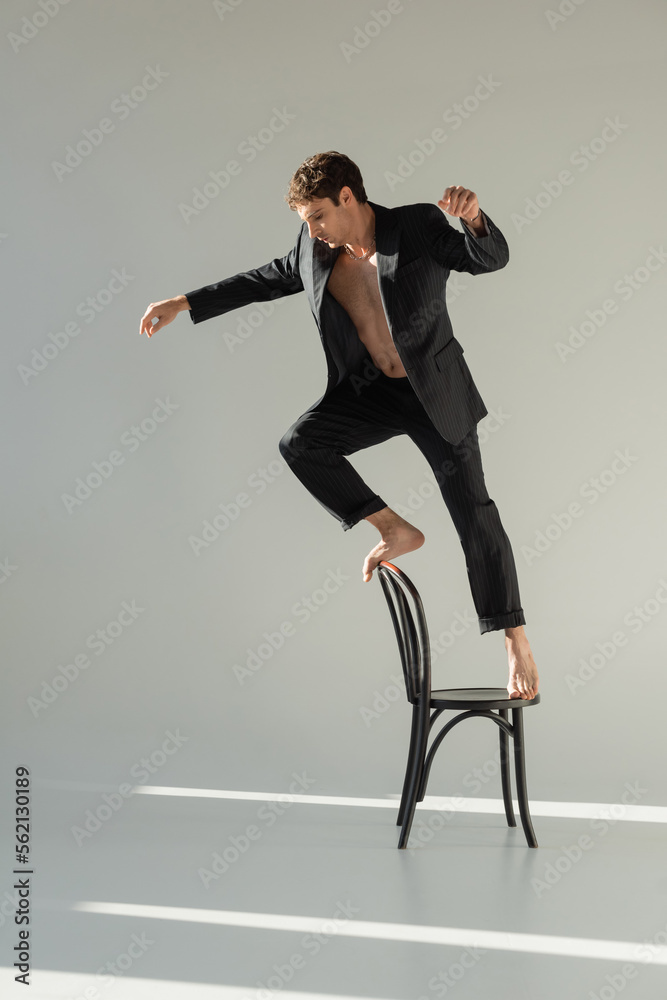 full length of barefoot and shirtless man in black suit doing trick while standing on chair on grey background.
