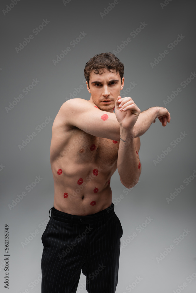 sexy shirtless man with red lipstick prints on body looking at camera while standing on grey background.