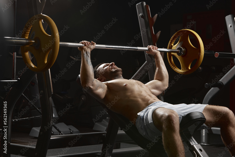 Portrait of young muscular man training shirtless in gym indoors. Doing bench press exercise. Endurance. Concept of sport, workout, strength - obrazy, fototapety, plakaty 
