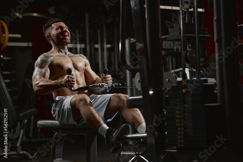 Portrait of young muscular man training shirtless in gym indoors. Pulling rope with weigh. Relief body shape. Concept of sport, workout, strength