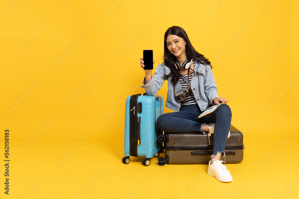 .Happy Asian woman traveler sitting on luggage and holding mobile phone isolated on yellow background, Tourist girl having cheerful holiday trip concept, Full body composition