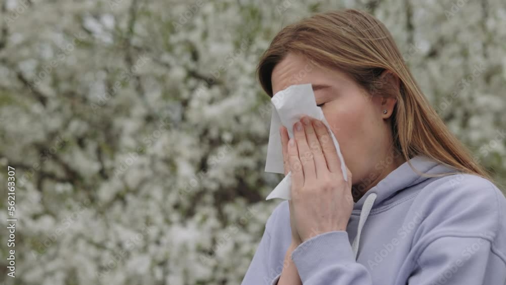 Unhealthy sick woman with allergy symptom blowing nose surrounded with seasonal flowering trees. Young caucasian lady having allergy symptoms outdoors Unhealthy sick woman with allergy symptom blowing nose surrounded with seasonal flowering trees. Young caucasian lady having allergy symptoms outdoors