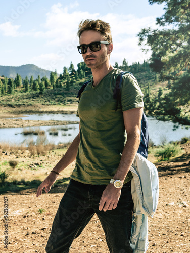 Male fashion model is hiking next to the lake by the mountains.